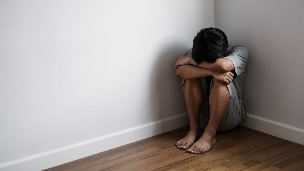 A young man curls up in a ball in a corner of a room