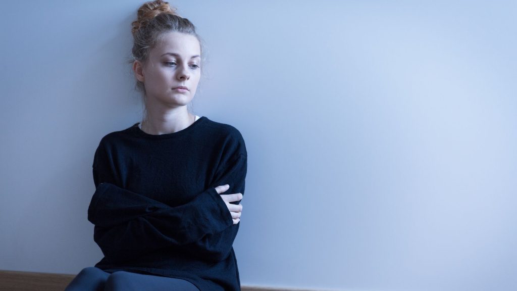 An uncomfortable looking woman in an undecorated room crosses her arms across her torso