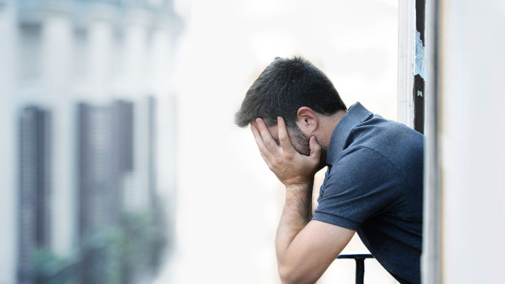 A man covers his face while his upper body extends out through a city apartment window
