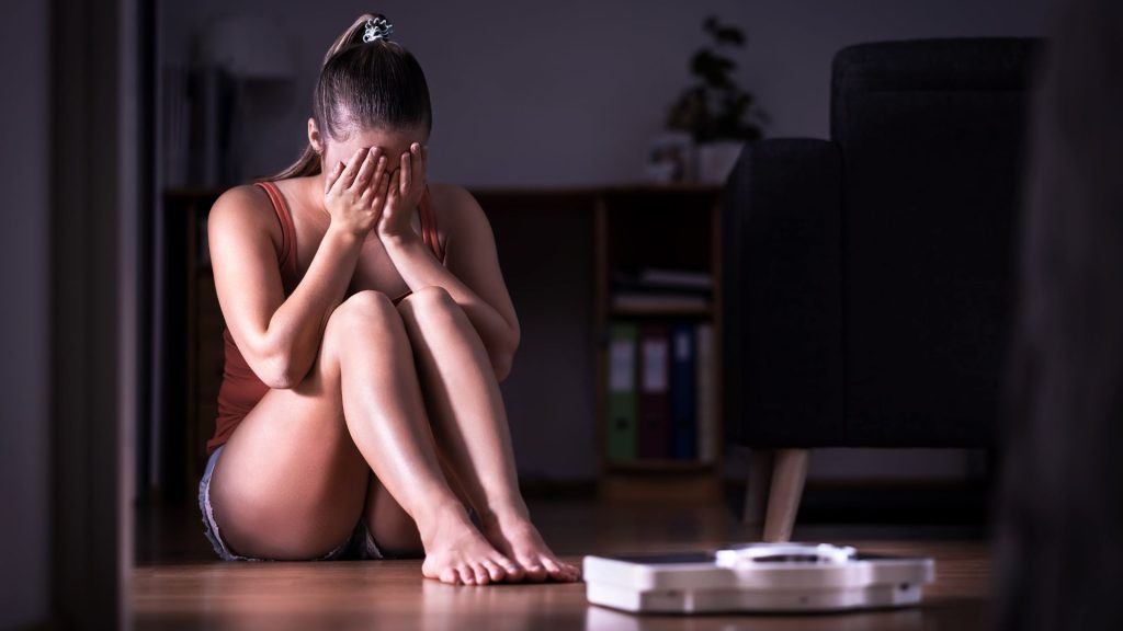 A woman looks distressed, covering her face, while sitting on a floor near a scale