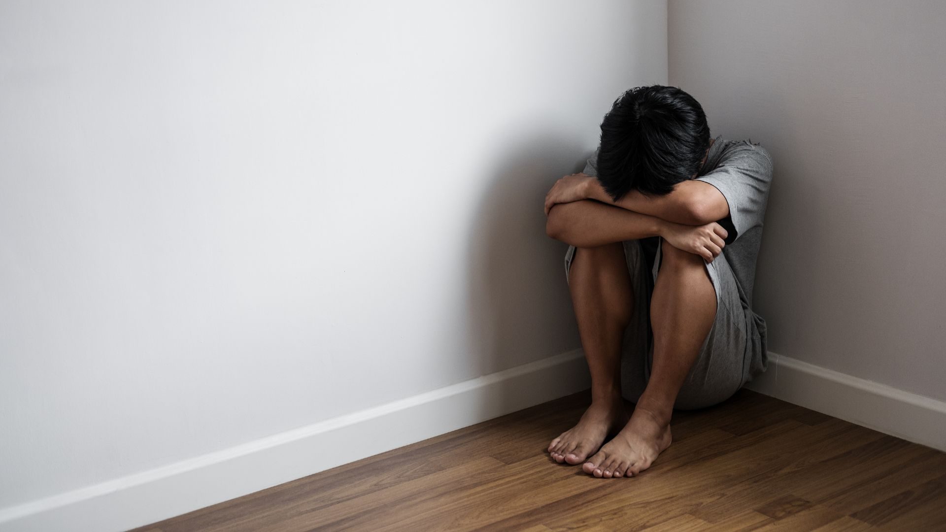 A young man curls up in a ball in a corner of a room