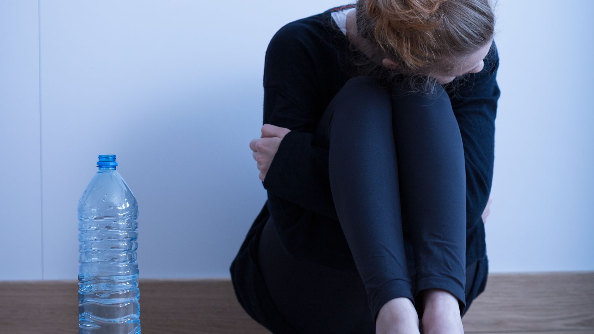 A woman holds her knees to her chest in a room with just a water bottle