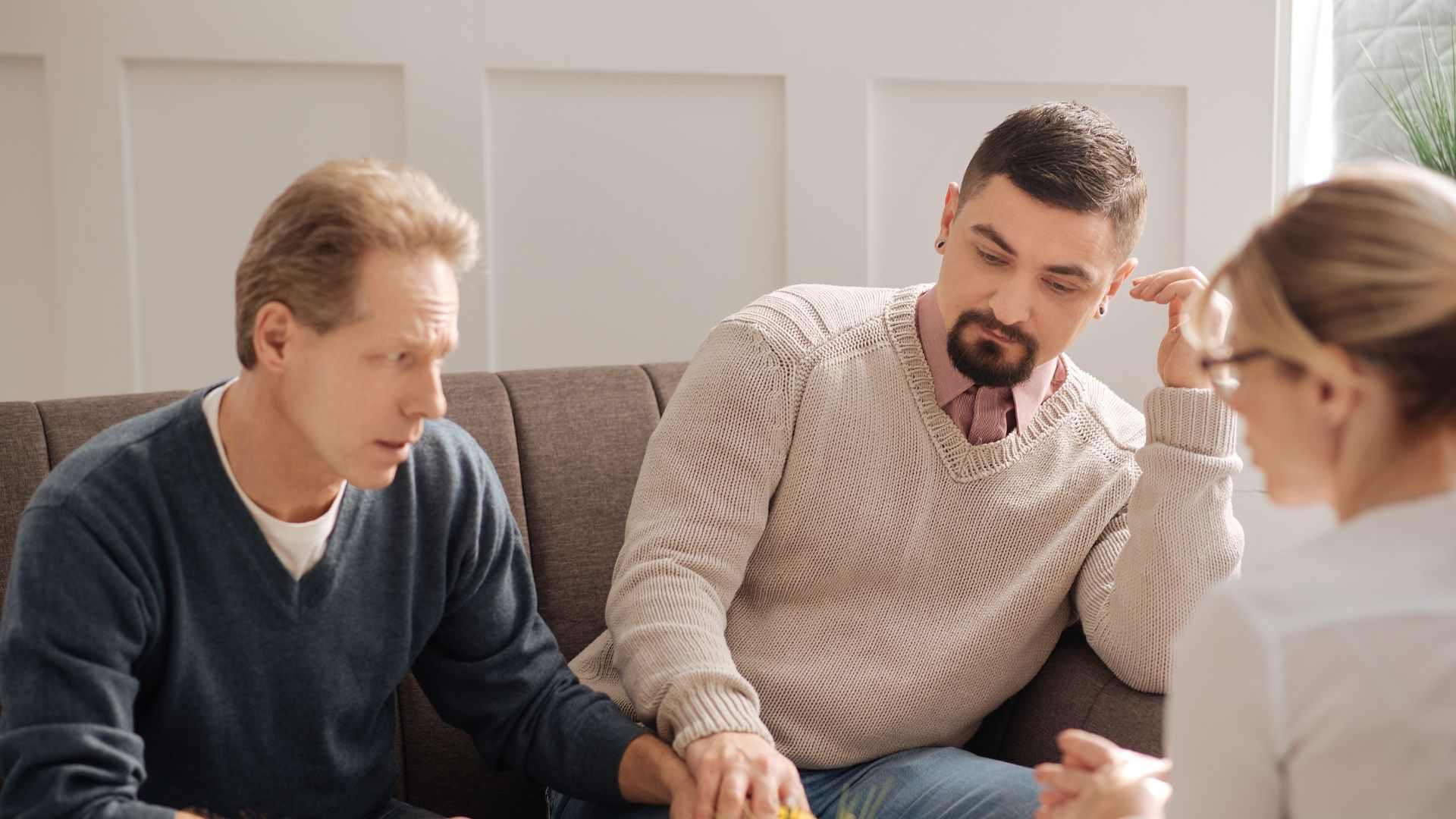 Two men hold hands, looking distressed, while sitting on a couch across from a therapist