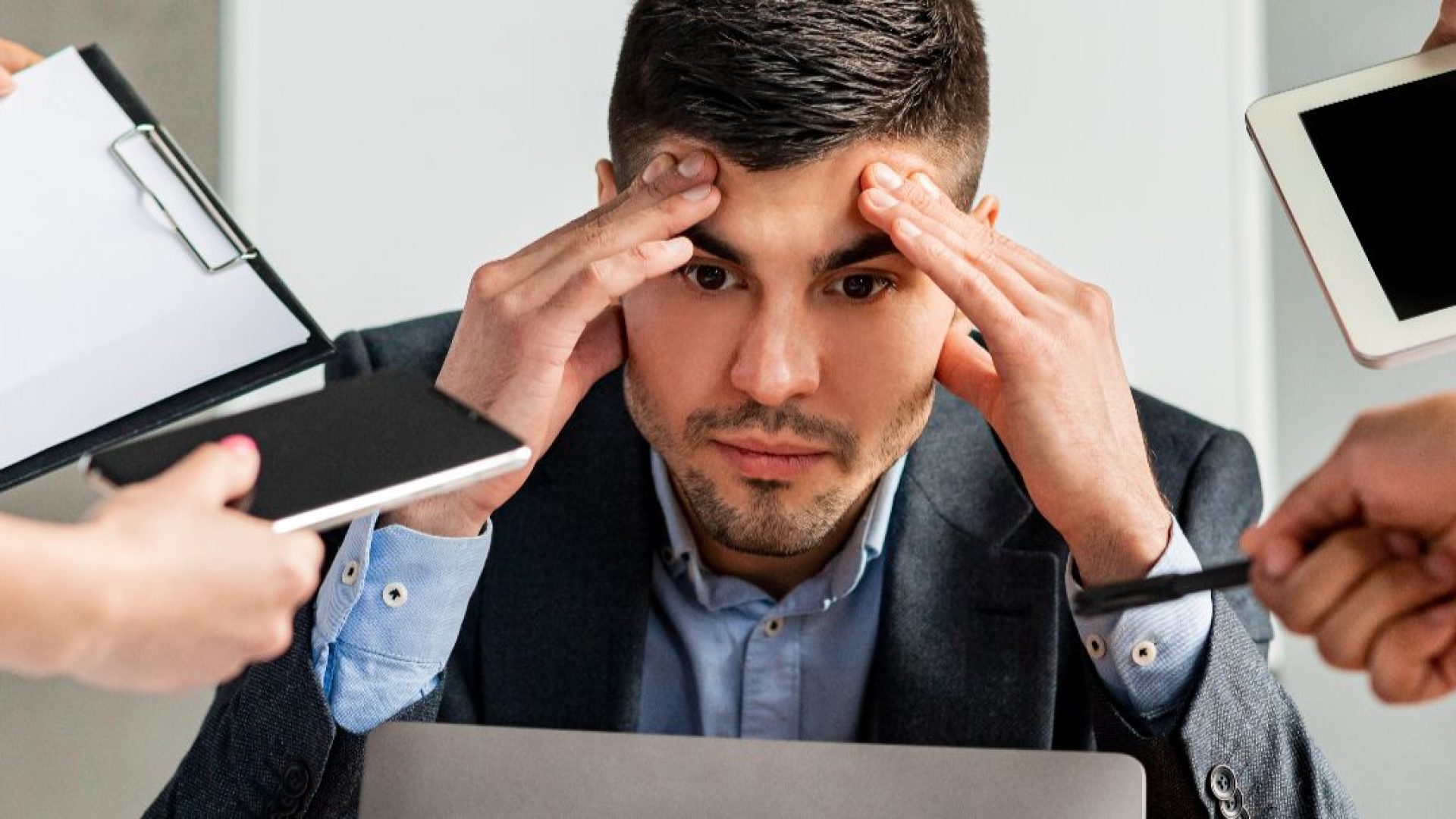 A stressed looking man surrounded by coworkers holding tablets, cell phones and paperwork