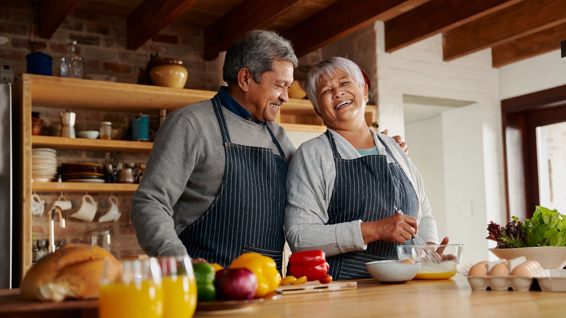 A happy looking older couple in a kitchen making eggs with peppers and bread also on the table