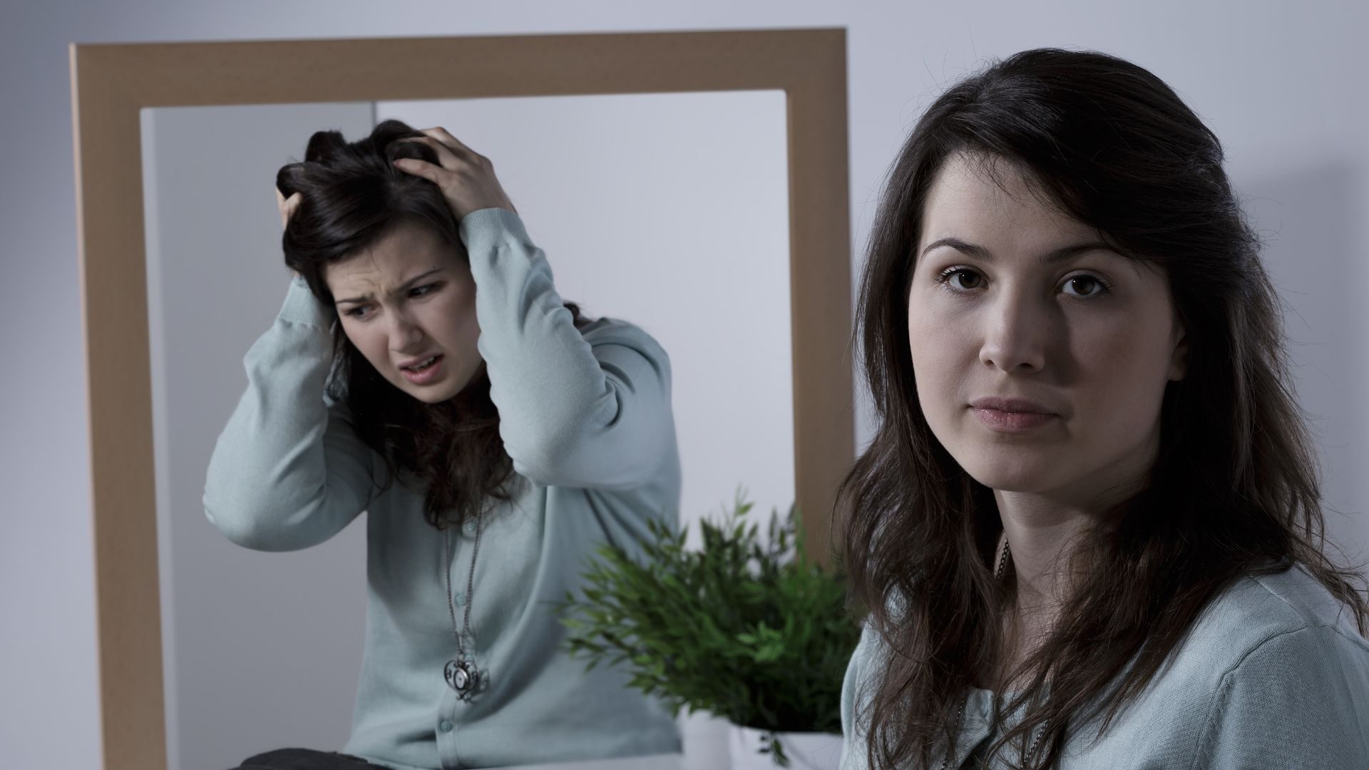 An image of a calm looking woman, but her reflection in the mirror looks distressed, with hands pulling her hair