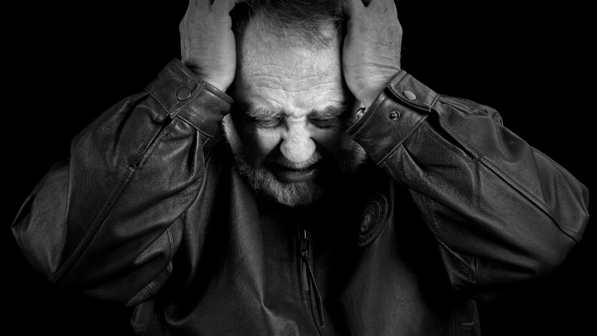 An older man in a leather jacket looks anguished, with his hands over his temples