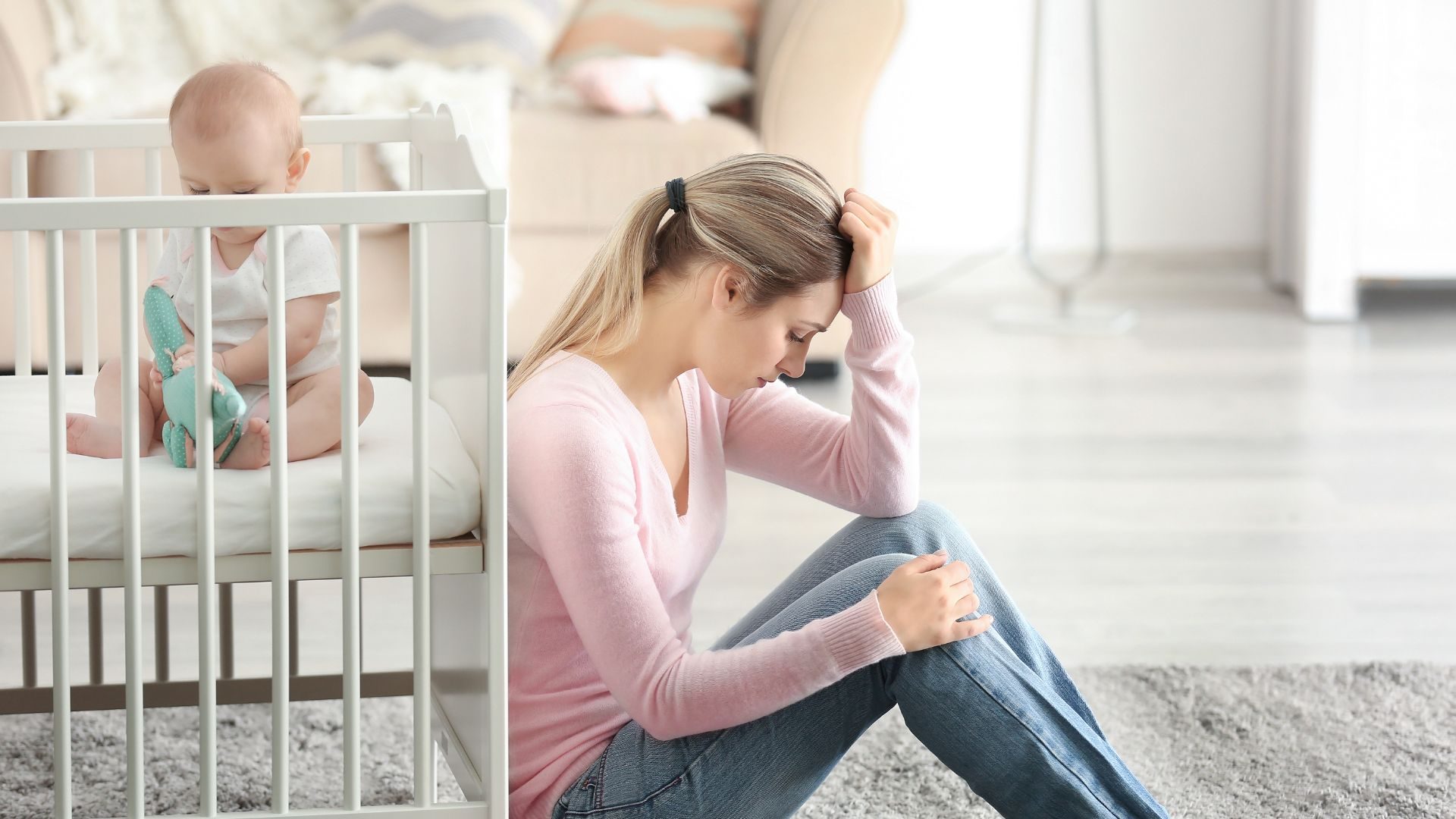 A new mother sits to the floor, hand against her head, while a baby plays in a crib