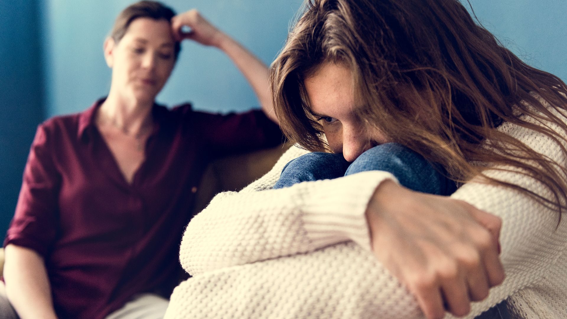 A young woman with her face against her knees, and arms around both, looks down while another older woman looks on uncomfortably