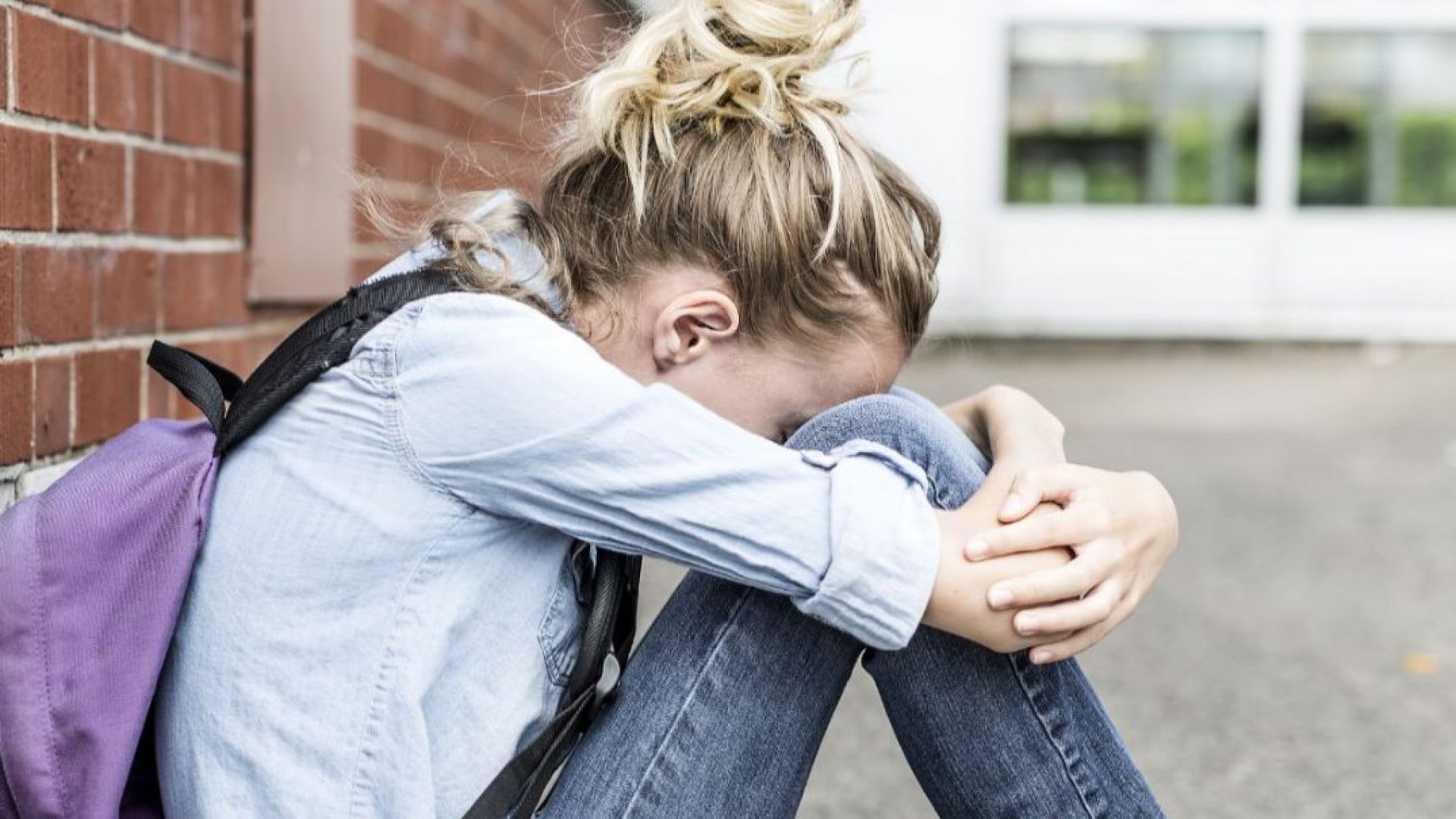 A teenager sitting against a wall outside a school, hiding their face.