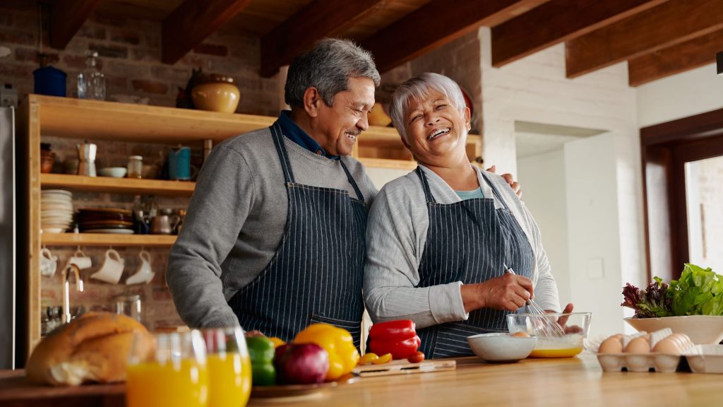 A happy looking older couple in a kitchen making eggs with peppers and bread also on the table