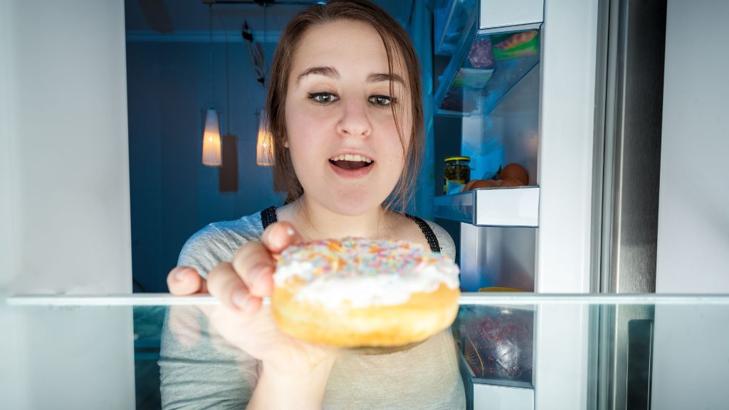A woman reaches into a refrigerator for a donut