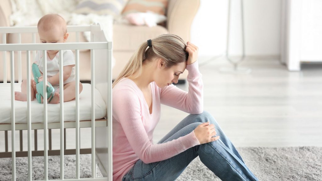 A new mother sits to the floor, hand against her head, while a baby plays in a crib