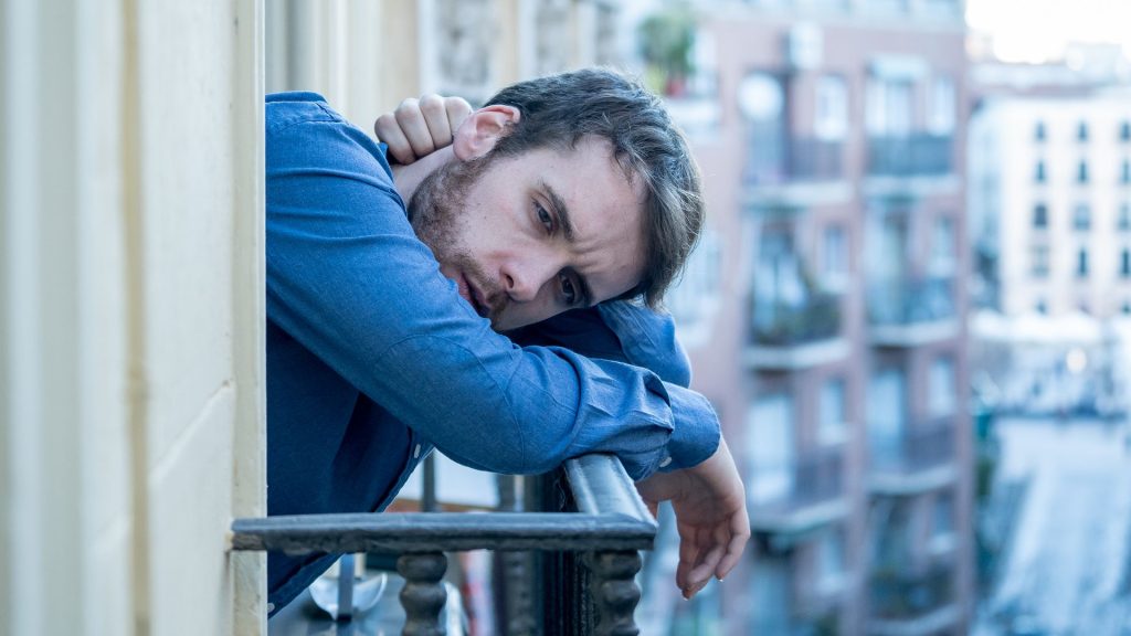 A man on a balcony looks out on a city street with an uncomfortable look on his face