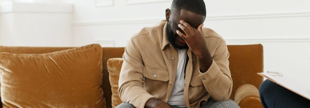 Person sitting on couch, holding hand to forehead, dealing with the symptoms of anxiety.