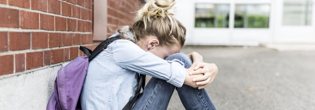 A teenager sitting against a wall outside a school, hiding their face.